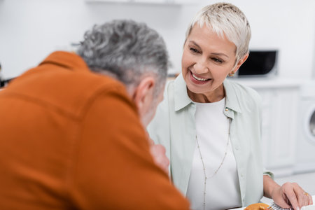 Cheerful senior woman looking at blurred husband near pancakes in kitchenの写真素材