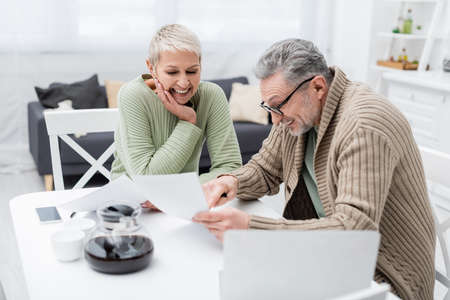 Positive pensioner pointing at paper near wife and devices in kitchenの写真素材