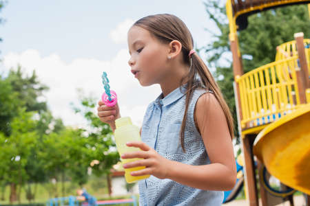 girl in sleeveless blouse blowing soap bubbles on playgroundの写真素材