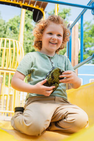 cheerful boy holding toy armored vehicle on slide in amusement parkの写真素材