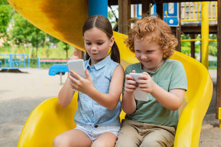 excited brother and sister sitting on slide and playing on smartphoneの写真素材