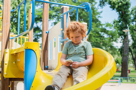redhead boy sitting on slide in amusement park and playing on smartphoneの写真素材