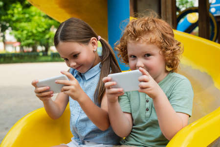 boy with mobile phone looking at camera near sister on playgroundの写真素材