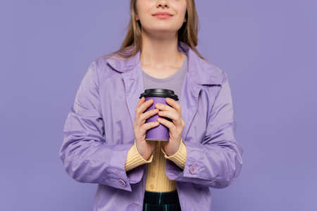 cropped view of smiling young woman in trench coat holding paper cup isolated on purple