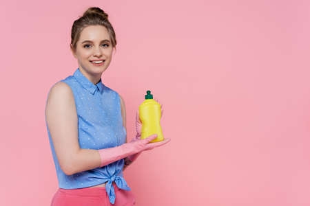 smiling young woman holding bottle with detergent isolated on pink