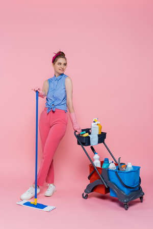 full length of smiling young woman in rubber gloves standing with mop near housekeeping cart on pink