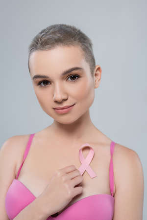 pretty young woman with breast cancer awareness ribbon looking at camera isolated on greyの写真素材