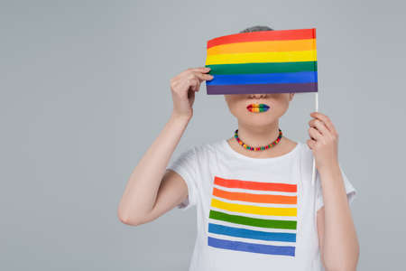 young woman in rainbow colors t-shirt obscuring face with lgbt flag isolated on greyの写真素材