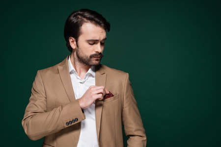 young man with mustache adjusting handkerchief in pocket of blazer on dark green