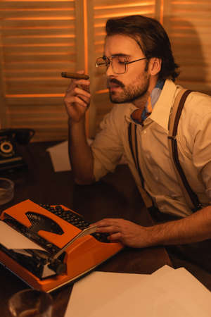 journalist with mustache and eyeglasses smoking cigar and looking at retro typewriter machineの写真素材