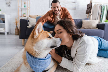 happy young woman cuddling akita inu dog near blurred boyfriend on couchの写真素材