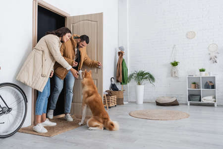 full length of happy couple in winter jackets standing near akita inu dog