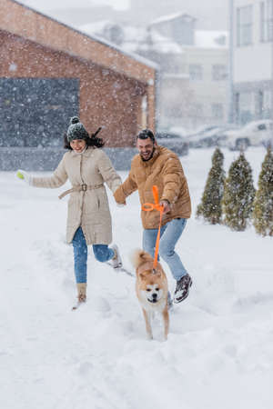 happy man holding leash while running together with girlfriend and akita inu dog under falling snowの写真素材