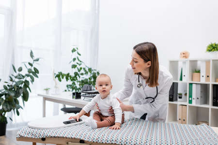 Smiling pediatrician holding baby near scales on medical couch in hospitalの写真素材