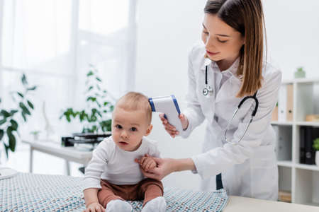 Baby boy looking at camera near family doctor with pyrometer in clinic