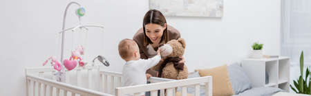 Young mother holding soft toy near baby boy in crib, bannerの写真素材