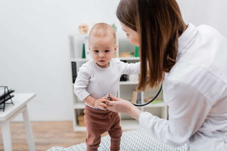Baby boy standing near blurred doctor with stethoscope on medical couch in clinicの写真素材