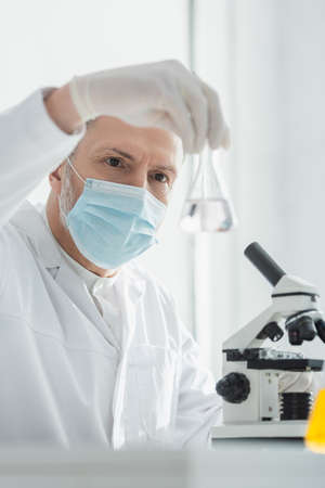 middle aged scientist in medical mask holding blurred flask near microscope