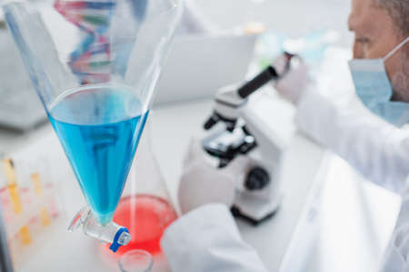 selective focus of flask with blue liquid near blurred scientist in medical mask
