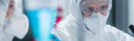scientist in hazmat suit, goggles and medical mask near blurred colleague in lab, bannerの写真素材