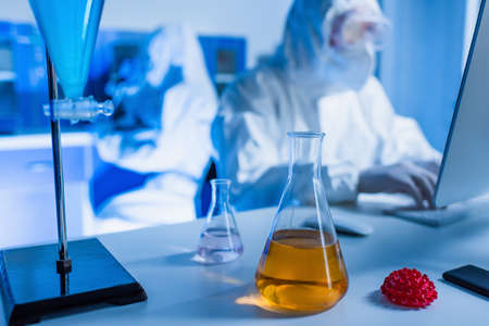 flasks with liquid and coronavirus bacteria model near blurred bioengineers in hazmat suitsの写真素材