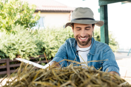 young bearded farmer in brim hat smiling near haystack on farmの写真素材