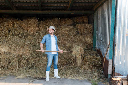full length view of happy farmer in denim clothes and rubber boots near haystackの写真素材