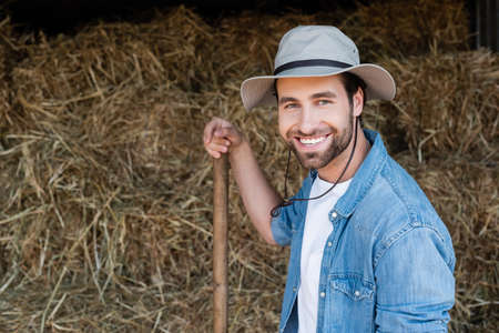 joyful farmer in brim hat looking at camera near blurred haystack on farmの写真素材