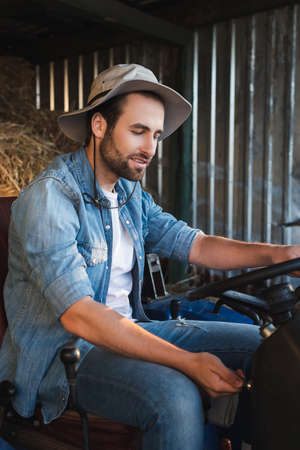 bearded farmer in brim hat and denim shirt sitting on tractor on farmの写真素材