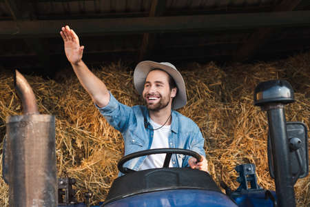 cheerful farmer on tractor looking away and waving handの写真素材