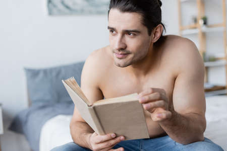dreamy and shirtless man with long hair holding book in bedroomの写真素材
