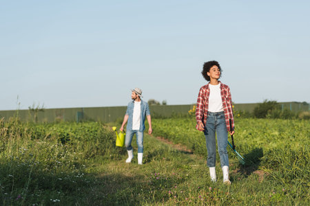 multiethnic farmers with rakes and watering can in farmland under blue skyの写真素材