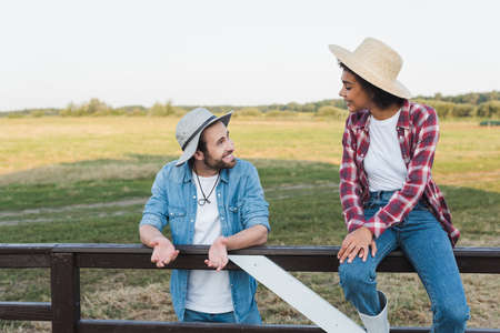 smiling farmer talking to african american colleague sitting on fence on farmland