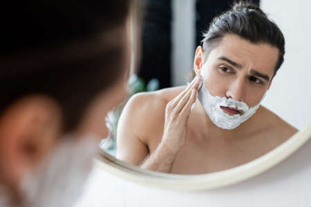 shirtless man applying shaving foam on face and looking at mirrorの写真素材