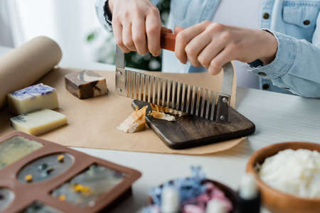 Cropped view of craftswoman cutting soap on board near blurred flowers at homeの写真素材