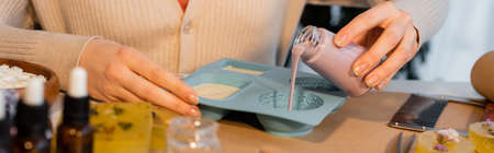 Cropped view of craftswoman pouring liquid soap in silicone mold near blurred essential oils, bannerの写真素材