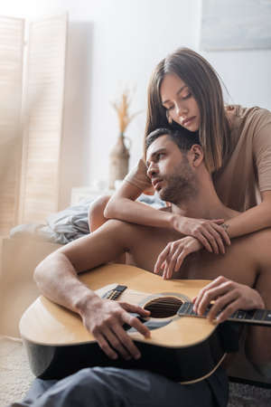 Brunette woman hugging shirtless man holding acoustic guitar in bedroomの写真素材