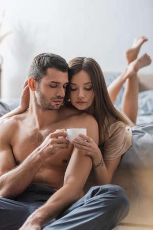Pretty woman holding cup of coffee near shirtless boyfriend in bedroomの写真素材