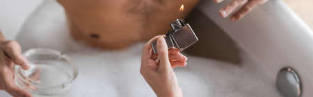 Cropped view of woman holding lighter near blurred man with ashtray in bathtub, bannerの写真素材