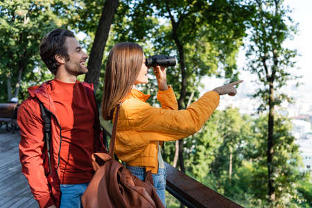 Side view of cheerful traveler looking through binoculars and pointing with finger near boyfriend...