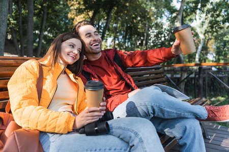Smiling tourist holding coffee to go and map near girlfriend with binoculars on bench outdoors