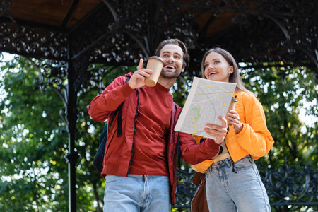 Low angle view of smiling tourist holding paper cup and map while pointing with finger near girlfriend outdoorsの写真素材