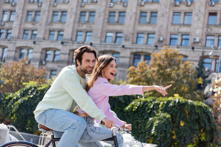 Excited woman pointing with finger while riding bike with boyfriend on urban streetの写真素材
