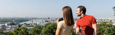 Positive tourist looking away near brunette girlfriend with binoculars on viewpoint in city, banner