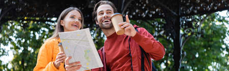 Low angle view of cheerful man holding map and pointing with finger near girlfriend with coffee to...
