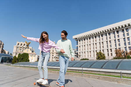 Happy woman riding penny board and holding hand of boyfriend on urban streetの写真素材
