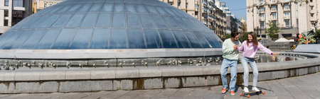 Positive couple riding penny board near building outdoors, bannerの写真素材