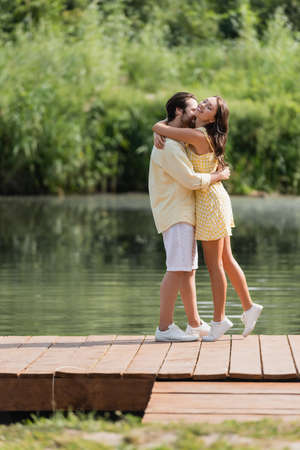 full length of joyful young couple in summer clothes hugging on pier near lakeの写真素材