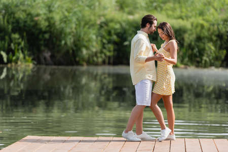 full length of happy young couple in summer clothes holding hands on pier near lakeの写真素材