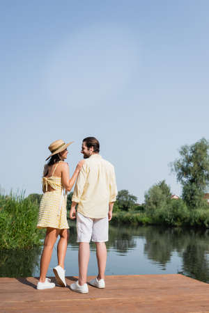 full length of happy young couple in summer clothes holding hands and standing on pier near lakeの写真素材